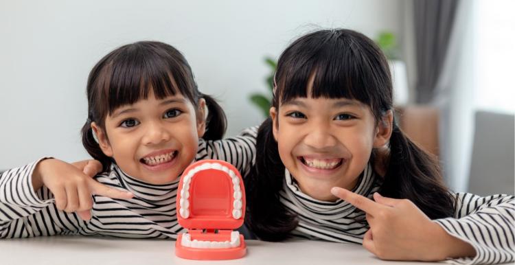 Two children smiling and pointing at their teeth behind a clicky teeth toy