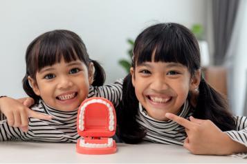 Two children smiling and pointing at their teeth behind a clicky teeth toy