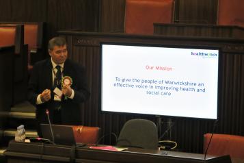 Chris Bain giving a presentation - next to a screen which says' Our mission - to give the people of Warwickshire an effective voice in improving health and social care.'